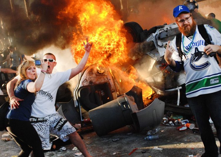 A riot surrounding a burning car. People look excited and are embracing chaos.