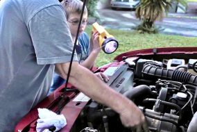 A son holds a flashlight for his father, who is working on a car.