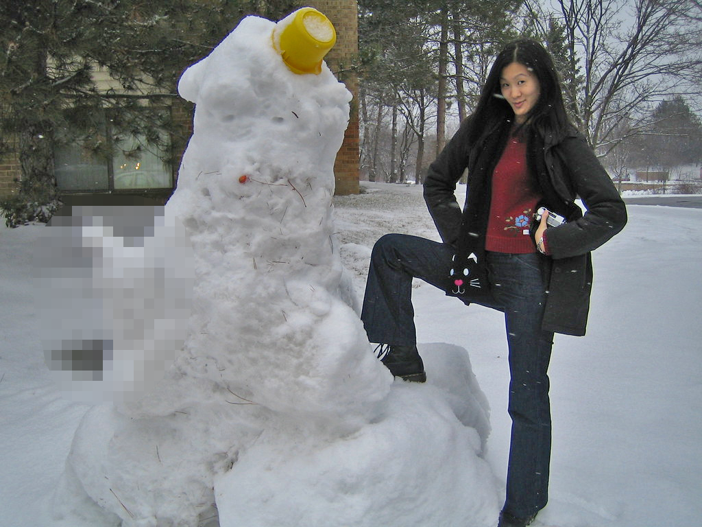 A large snowman with comically large and censored genitalia, and a woman proudly posing next to it.