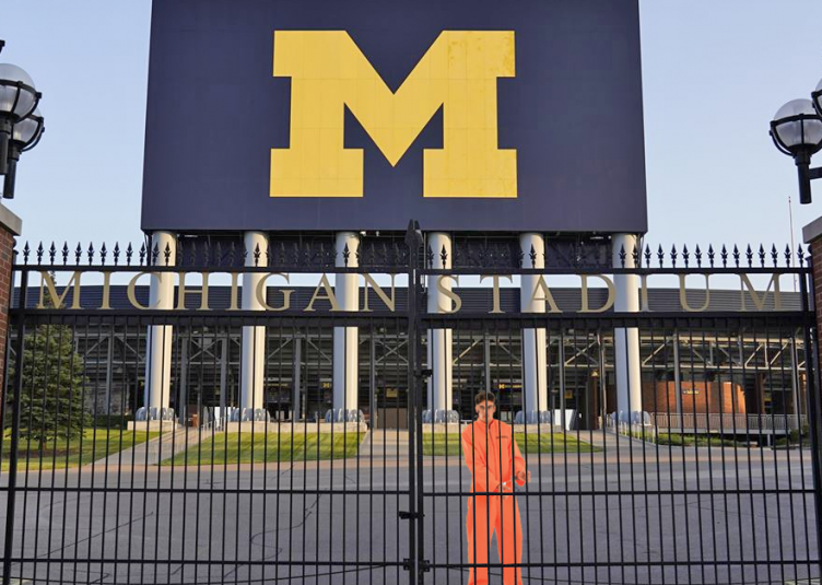 A man in an orange jumpsuit behind the gates of the Ann Arbor Football Stadium.