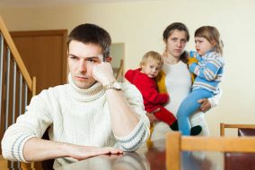 Man sitting disgruntled at a dining table with a woman behind him holding some small children.