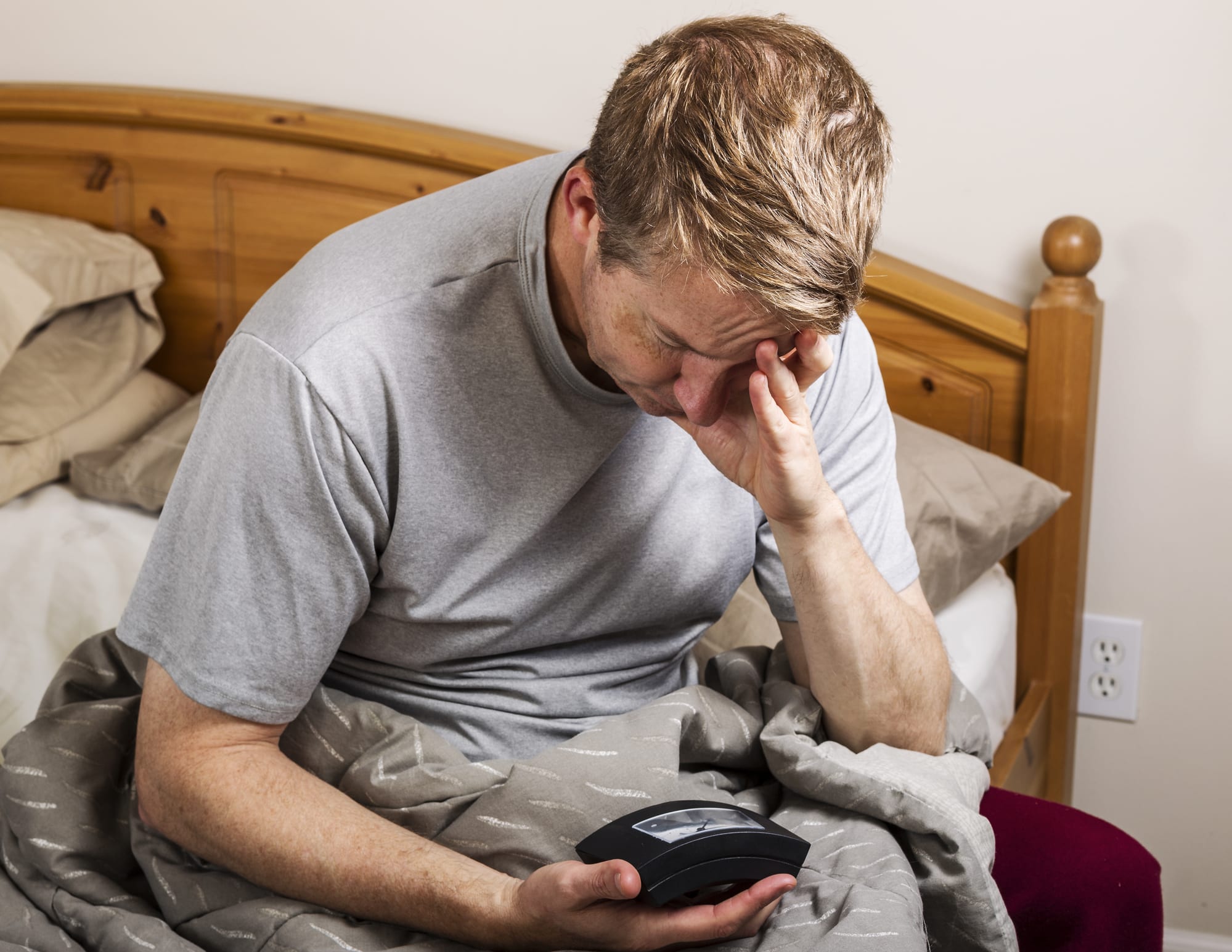 Blond man sitting in bed, looking tiredly at alarm clock