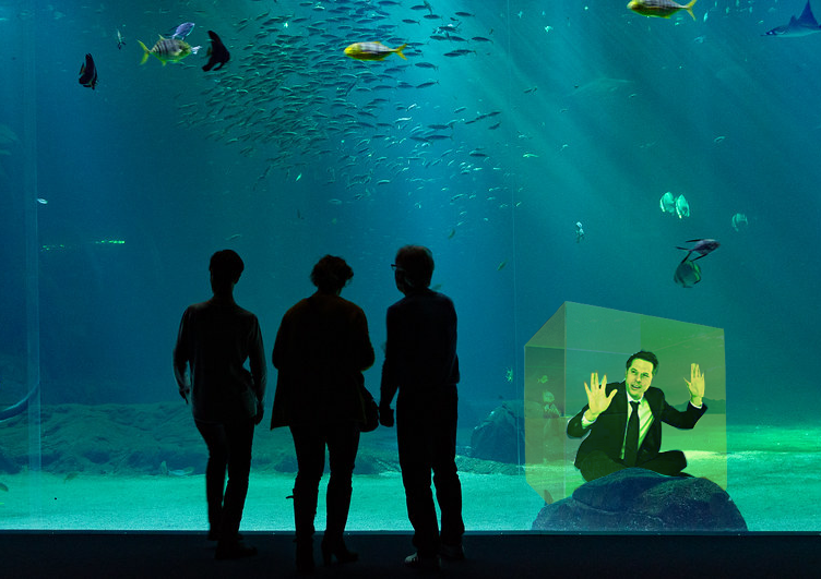 Picture of a family looking at a large tank that has fish and a man trapped inside of a box.