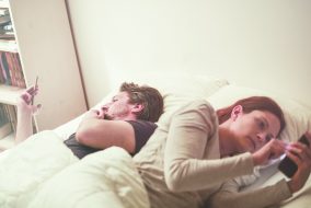 Two people lying in bed, both looking at their phones uninterested