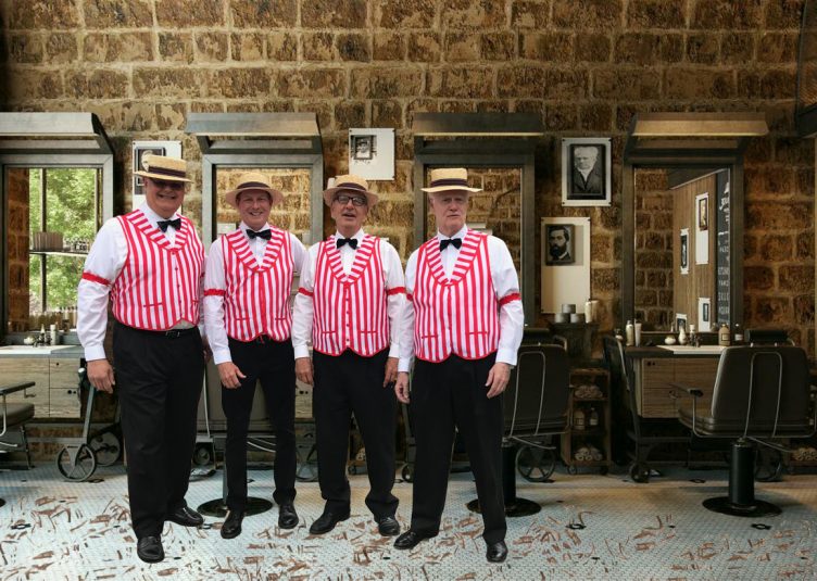 4 members of a barbershop quartet, in the striped jackets and odd hats, are performing in an actual barber shop
