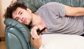 A man lies on a couch holding a TV remote.