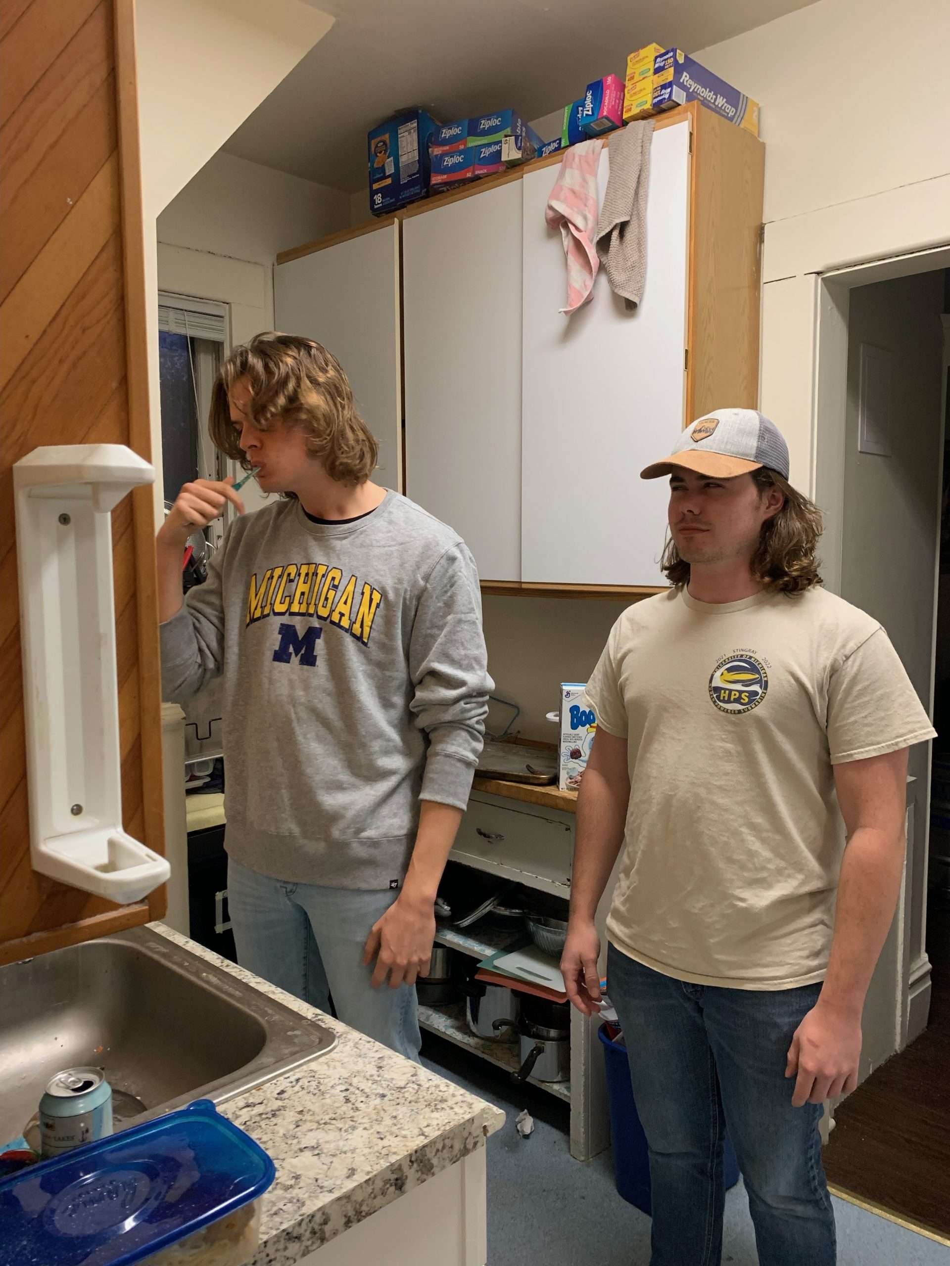 Two men standing at a sink, one brushing his teeth.