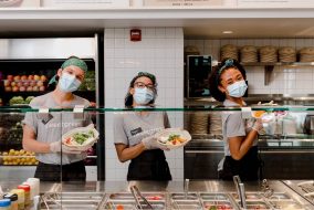 Three Sweetgreen workers stand behind the counter and hold up bowls of food.