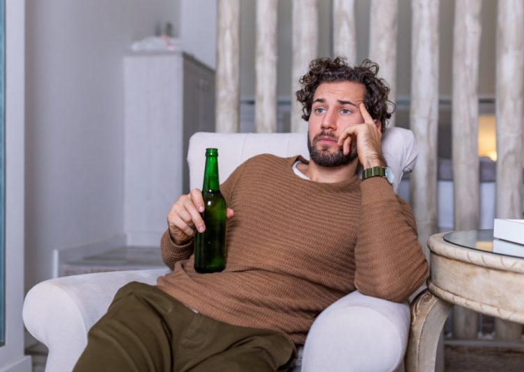 A sad looking man holding a beer staring whistfully at the TV