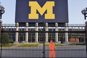 A man in an orange jumpsuit behind the gates of the Ann Arbor Football Stadium.