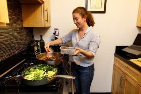 A woman is cooking meat in veggies over a stovetop in a small apartment.
