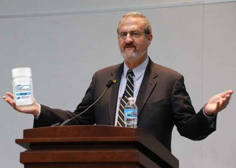 Mark Schlissel edited to holding container of Viagra while standing at podium