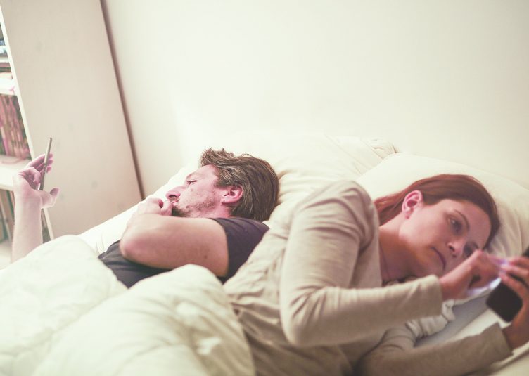 Two people lying in bed, both looking at their phones uninterested