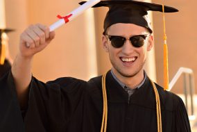 Happy man in graduation hat and robe holding up degree