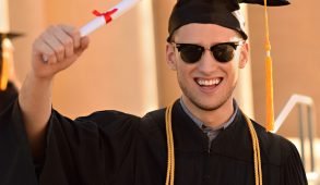 Happy man in graduation hat and robe holding up degree