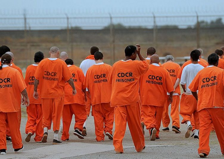 Prisoners in orange uniforms walk towards a barbed-wire fence.