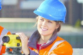 A smiling woman wearing a blue hardhat and orange safety vest holds a piece of equipment