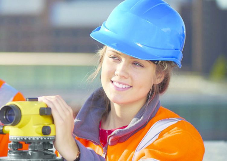 A smiling woman wearing a blue hardhat and orange safety vest holds a piece of equipment