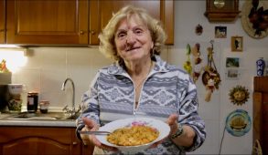 A older woman holds up a bowl of food to the camera.