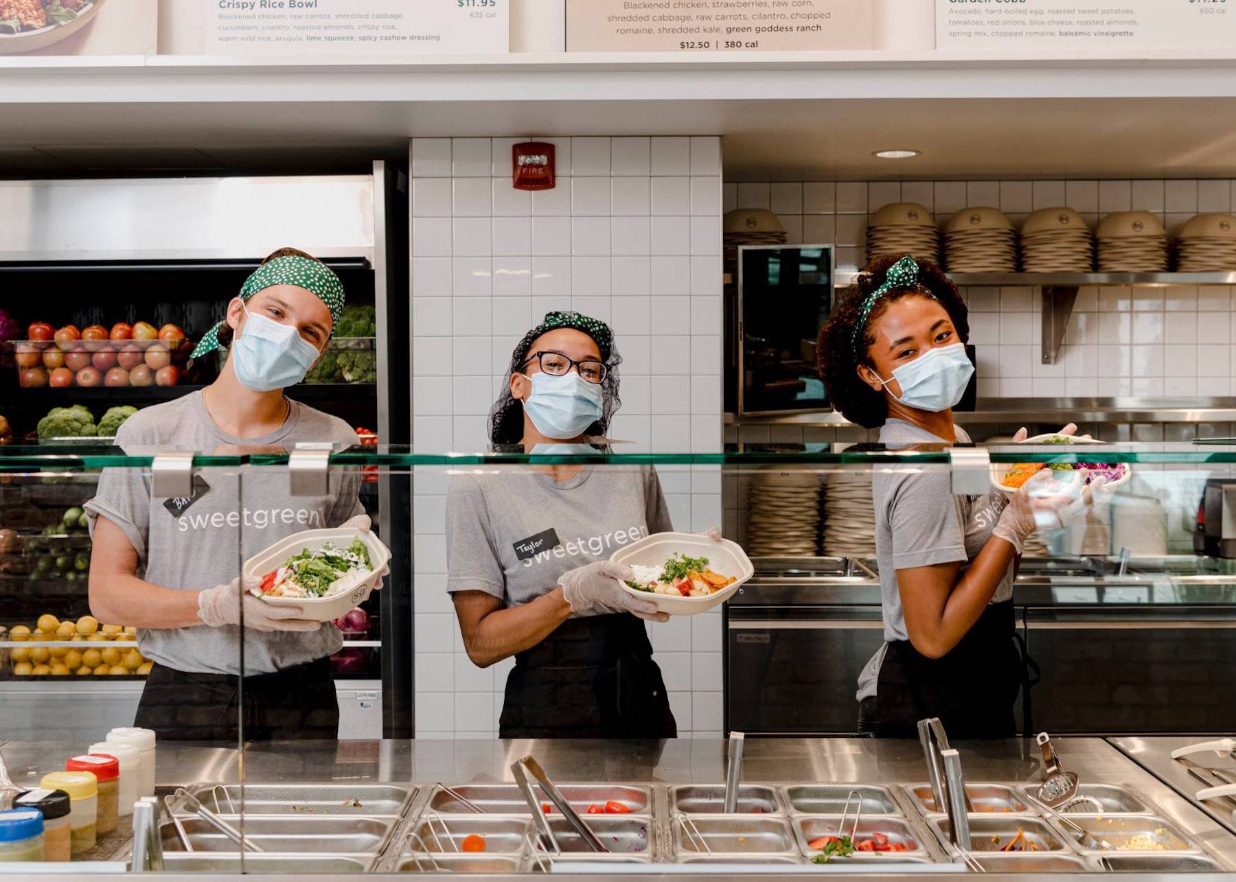 Three Sweetgreen workers stand behind the counter and hold up bowls of food.