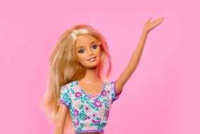 A barbie wearing a floral top waves in front of a pink background.