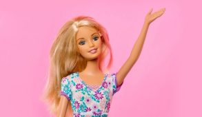 A barbie wearing a floral top waves in front of a pink background.