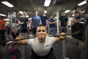 A man wearing a grey shirt lifts at a squat rack.