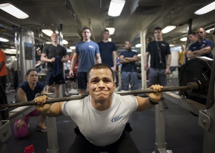 A man wearing a grey shirt lifts at a squat rack.