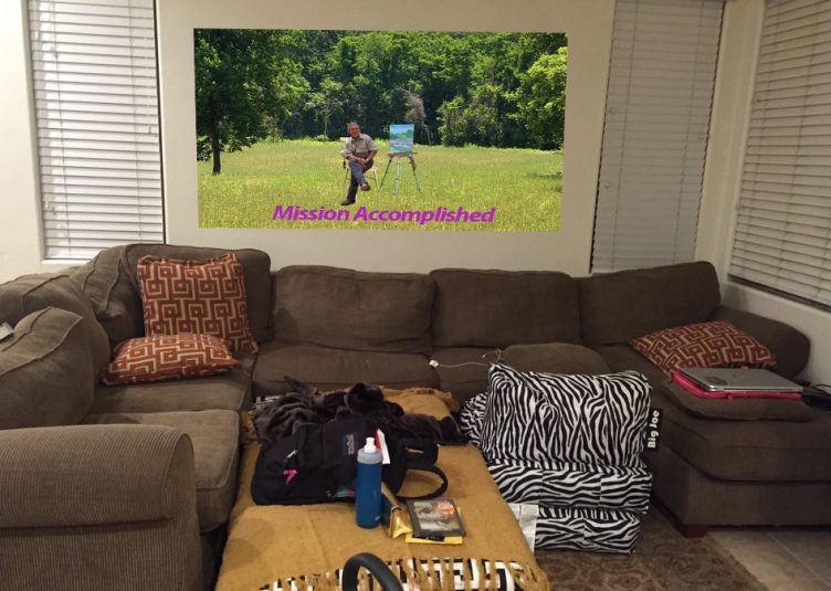 A banner of George W Bush painting in a field with the text "Mission Accomplished," hanging in a college living room.